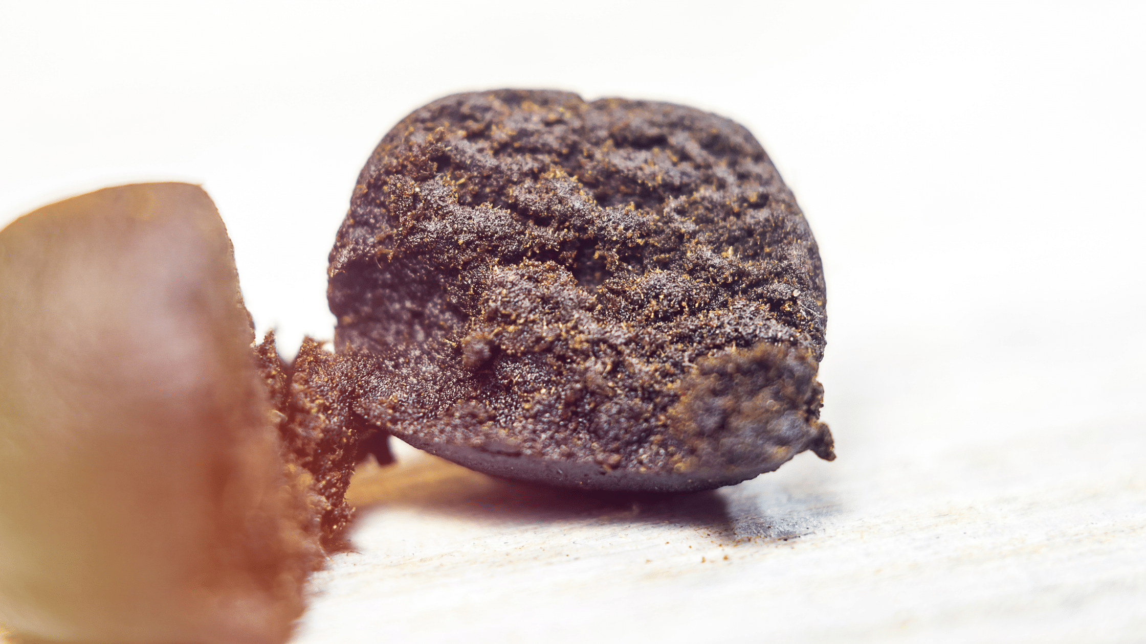 Close-up of a ball of hand-rubbed cannabis resin held between fingertips on a pale wooden surface, showing the crumbly, oily brown texture of fresh charas.