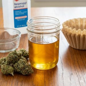 A mason jar filled with golden honey oil sitting on a wooden table next to dried cannabis buds, unbleached coffee filters, and a bottle of isopropyl alcohol.