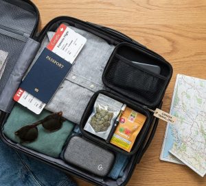An open suitcase on a wooden floor containing a Canadian passport, airline ticket, map, and a specialized smell-proof case packed with cannabis flower, a vape pen, and gummies.